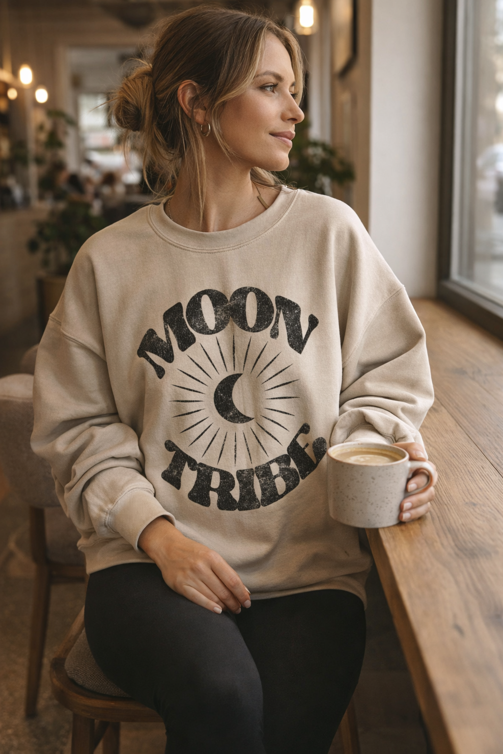 Contemplative woman wearing a Fitkitty Culture Lunar Essence Fleece Crew with moon tribe graphic sweatshirt in a cozy coffee shop setting