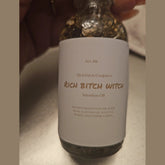 Rich Bitch Conjure Hoodoo Oil bottle labeled "Rich Witch Bitch" with dark, mystical packaging and a wooden surface background.