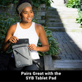 Woman sitting outside, placing a tablet pad into a Shield Your Body SYB Sling Bag, a stylish backpack for everyday use.