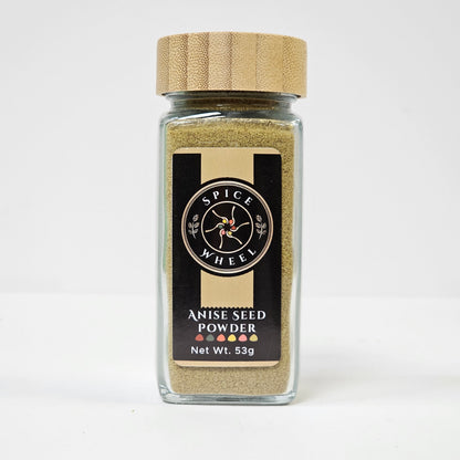 Fine greenish-brown anise seed powder in a clear glass jar from Spice Wheel, showing its smooth texture and aromatic quality.